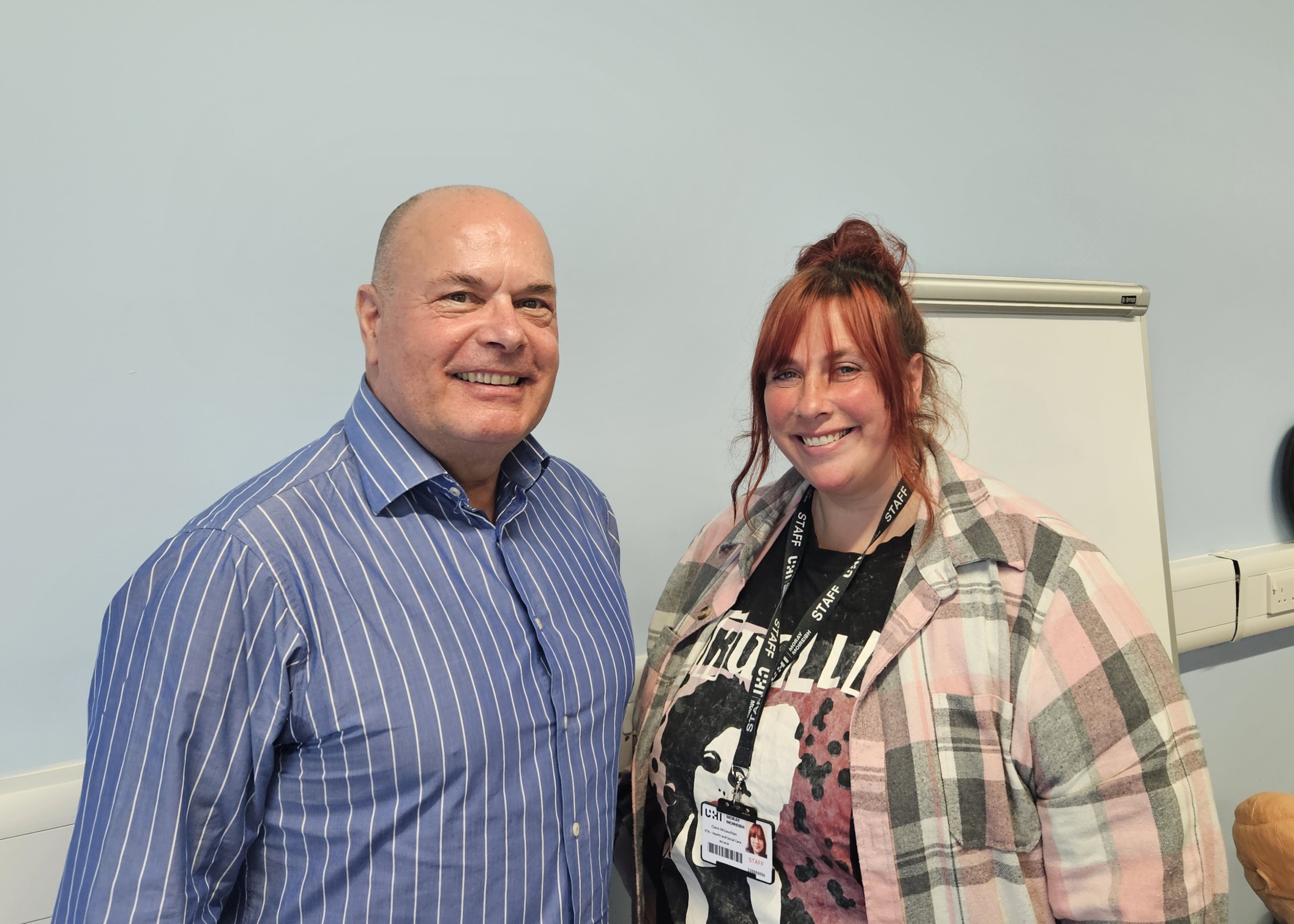 Photograph of UHI Moray's First Aid Trainers, Mike and Claire. They are stood side by side in front of a pale blue wall, smiling at the camera.